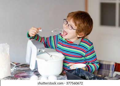 Beautiful Funny Blond Little Kid Boy N Glasses Baking Cake And Tasting Dough In Domestic Kitchen. Happy Child Having Fun With Working With Mixer, Flour, Eggs And Fruits At Home.