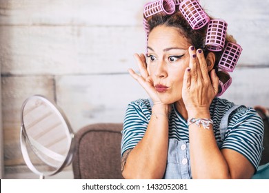 Beautiful And Funny Expression Caucasian Adult Woman Getting Ready At Home In Front Of The Mirror With Make Up On The Face - Wrinkles Checking And Get Older Concept For Young People - Curlers On Hair