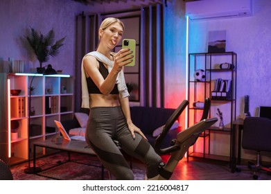 Beautiful Funny Smiling Young Woman Showing Tongue While Taking Selfie On Her Smartphone While Doing Cardio On Stationary Bike. Attractive Female Working Out On Fitness, Training On Exercise Bike.