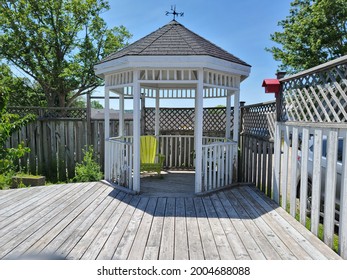 A Beautiful Gazebo Tucked Into The Corner Of A Backyard, Deck Made Of Wood. The Deck Is Surrounded With A Wooden Gate With A Lattice Design. There Is A Weathervane On Top Of The Gazebo And A Birdhouse