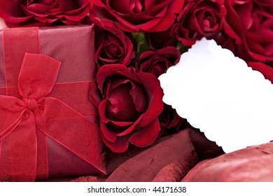 Beautiful Gift Box With A Dozen Red Roses And Blank Card Lying On A Two Toned Red Background. Extreme Shallow DOF With Focus On Gift And Center Roses.