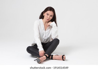 A beautiful girl of 25 years old, European appearance, with dark long hair, in a white shirt and black trousers, sits in a light studio and looks mysteriously. Pleasure, relaxation, availability. 