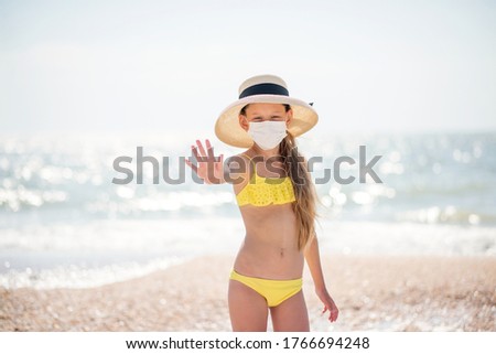 Beautiful girl alone on the beach in a medical mask. A crisis. How to wear a mask when traveling. Family content. Child isolated on the beach. Keep your distance. Beautiful long-haired girl in a swims