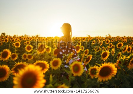 Beautiful girl with the American flag in a sunflower field. 4th of July. Fourth of July. Freedom. Sunset light The girl smiles. Beautiful sunset. Independence Day. Patriotic holiday. 
