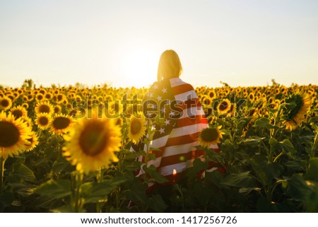 Beautiful girl with the American flag in a sunflower field. 4th of July. Fourth of July. Freedom. Sunset light The girl smiles. Beautiful sunset. Independence Day. Patriotic holiday. 