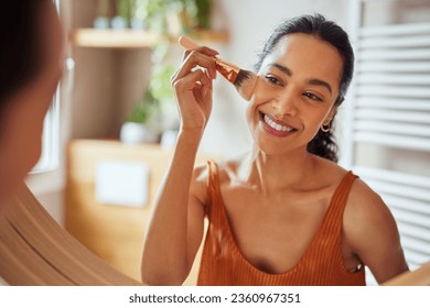 Beautiful girl applying makeup using powder brush before going to work. Healthy latin woman looking in the mirror and applying cosmetic with a big brush. Young woman applying foundation or blusher.
