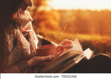 Beautiful girl in autumn forest reading a book covered with a warm blanket.a woman sits near a tree in an autumn forest and holds a book and a cup with a hot drink in her hands. Girl reading a book