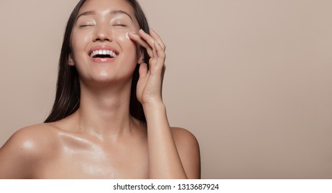 Beautiful girl with bare shoulders applying cream on her face and smiling against beige background. Smiling asian woman with glowing skin applying facial skincare cream with eyes closed.