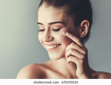 Beautiful Girl With Bare Shoulders Is Applying Cream On Her Face And Smiling, On Gray Background