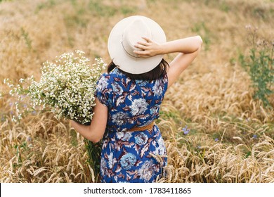Beautiful girl with big daisies bouquet, back view. Tranquil summer in countryside. Stylish young woman in blue vintage dress and hat posing with white wildflowers in wheat field meadow.