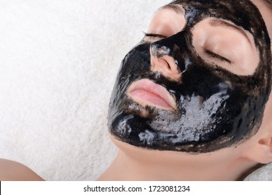 A beautiful girl with a black mask on her face is lying with her eyes closed on a white soft towel. Black face mask with activated carbon. Face care in the salon or at home, cleaning the pores.
