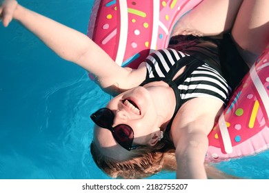 Beautiful girl in a black and white swimsuit and sunglasses enjoys relaxing in the pool on a hot sunny day. Floats in the pool on an inflatable circle in the shape of a donut, smiles and has fun