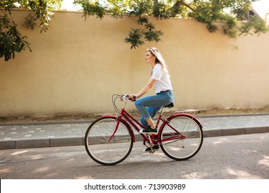 Beautiful girl with blond hair happily riding bicycle. Portrait of young lady in jeans and sunglasses on head having fun while riding on red bicycle along city streets