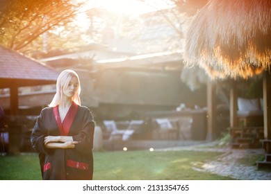 A beautiful girl with blonde hair in a traditional Chinese black and red dress. Portrait of an attractive woman against the background of wooden buildings in Asian style. Sunset in a green park.