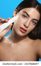 Beautiful girl with blue eyes, has bare shoulders, shows tube of lotion or cream for face, skin care routine spa cosmetic, applies moisturizer, stands over blue background