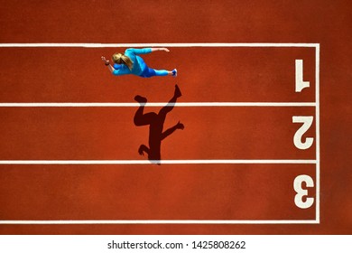 Beautiful Girl In A Blue Sportswear And Sneakers Starts Her Sprint Run With A Long Jump On The Running Track Number 1 At The Stadium Outdoors. Sun Shines Onto Her Body. Top View Aerial Photo.