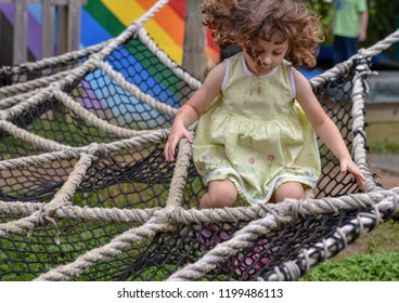 A Beautiful Girl With Bouncing Curls - Hair That Defies Gravity By Flying Above Her Head - Jumps In An Oversized Spider Web Woven From Ropes At An Outdoor Playground.