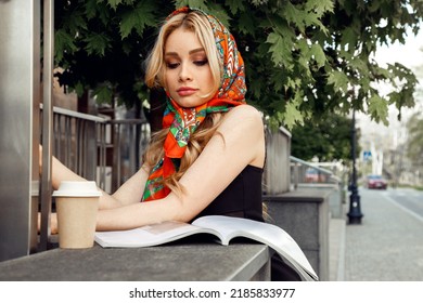 Beautiful Girl With Bright Makeup In Stylish Colorful Headscarf Looking Down And Reading Magazine With Glass Of Coffee In Morning Closeup. Paris Style, Cityscape, Urban Lifestyle, Street Walking. 