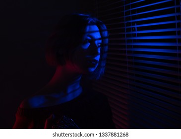 A beautiful girl with a chain in her hands stands near the window with blinds in the light of neon lights of blue and red light - Powered by Shutterstock