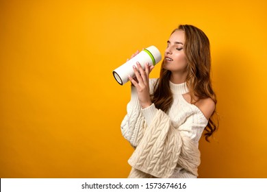 Beautiful Girl With Clean Skin And Loose Curls In A White Sweater On A Yellow Background, Brings A Thermo Mug For A Sip