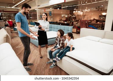 Beautiful girl consultant demonstrates orthopedic mattress to confident man with children in furniture store. Purchase of orthopedic mattress. Sale of orthopedic mattress in furniture store.