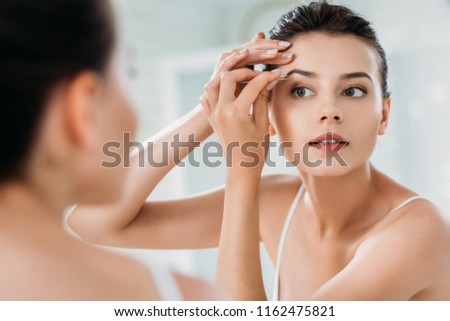 beautiful girl correcting eyebrows with tweezers and looking at mirror in bathroom 