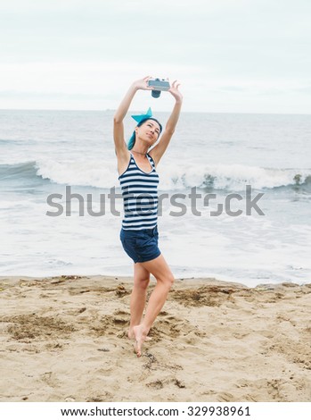 Beautiful girl in costume of sailor and paper ship in hairstyle doing selfie with vintage photo camera on beach in summer, pin-up image
