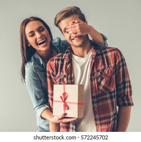 Beautiful girl is covering her boyfriend eyes and giving him a present, on gray background - Shutterstock ID 572245702