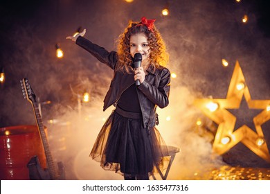 Beautiful Girl With Curly Hair Wearing Leather Jacket, Boots Sing Into A Wireless Microphone For Karaoke Like Rock Star In Recording Studio Or Stage. Smoke On Background.