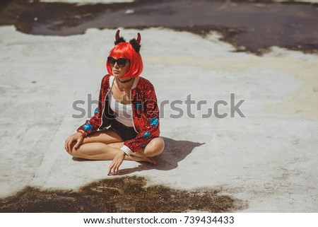 Beautiful girl devil sitting on city background near the puddle with his reflection. In anticipation of Halloween. Portrait. Wears black sunglasses