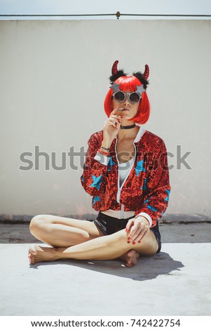 Beautiful girl devil sitting on white background and smoke cigar. In anticipation of Halloween. Portrait. Wears black sunglasses