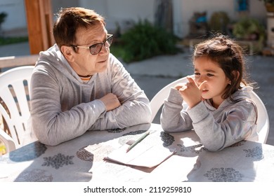 Beautiful Girl Doing Homework With The Help Of Her Grandmother On An Outdoor Patio.Close Up Portrait Of Senior Teacher Supervising Little Kid Doing Homework Back Yard.Young Child Counting On Fingers.