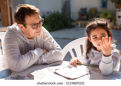 Beautiful Girl Doing Homework With The Help Of Her Grandmother On An Outdoor Patio.Close Up Portrait Of Senior Teacher Supervising Little Kid Doing Homework Back Yard. Young Child Counting On Fingers.