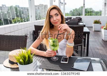 Beautiful girl dreams sitting in cafe, with long hair summer veranda. On tablet, glass lime cocktail phone straw hat table. Emotions fantasy thoughts, experiences and memories, thinks and decides.