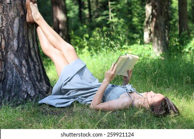 Beautiful girl in dress lying under tree on grass and reading book