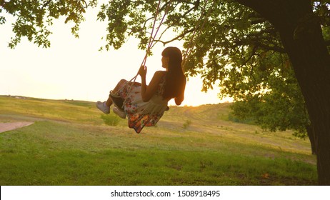 Beautiful Girl In A Dress In A Park On A Swing Flies. Young Girl Swinging On A Rope Swing On An Oak Branch. Teenage Girl Enjoys Flying Summer Evening In Forest