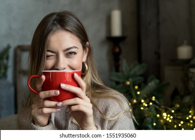 Beautiful Girl Drinking Coffee Or Tea From A Red Cup And And Winks. Christmas Mood With Copy Space