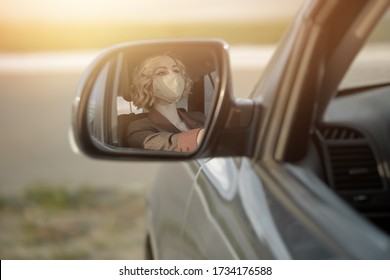 Beautiful Girl Driver With A Mask On Her Face, Sitting At The Wheel Of The Car Reflected In The Side Mirror Of The Car At Sunset.