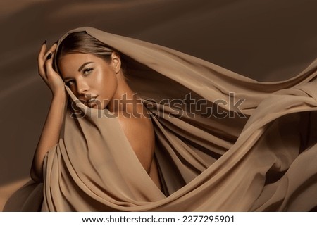 Beautiful Girl Face with Natural Makeup and Sun Tanned Skin. Beauty Woman wrapped in Brown Silk Fabric waving on Wind. Fashion Model Portrait wearing Head Satin Scarf