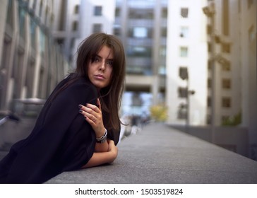 Beautiful girl fashion portrait. Leaning on staircase in black classic jacket over the shoulders. Holding one shoulder with left hand. Looking away.