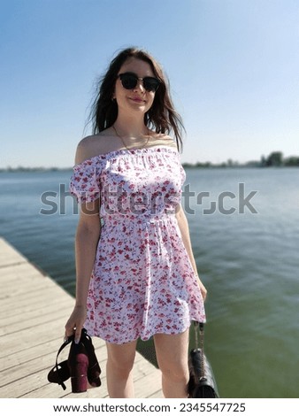 Beautiful girl in a flowered summer dress and sunglasses stands near the water and holds burgundy sandals in her hands