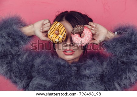 Beautiful girl in a fur coat with donuts in hands posing on a pink background.