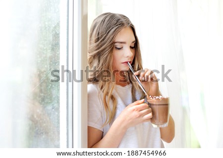 Beautiful girl with a glass cup of cocoa and marshmallows drinks with a straw in hands near window at rainy day