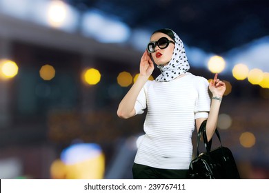 Beautiful Girl With Gold Lights Background. Luxury, Vintage Girl With A Bag Walk Around The Store. Woman In Sunglasses And A Scarf With A Female Handbag Shopping. Model Girl In Vintage Retro Style.