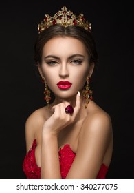 Beautiful girl in a golden crown and earrings on a dark background. Ring in the heart shape. Red dress and red lipstick.