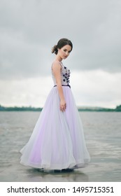 Beautiful Girl In Graduate Dress Near Water. Young Girl At The Prom With Gorgeous Hair In Tender Purple Dress. Professional Make-up And Hairstyle, Stylish. Alone Girl Stands On A Pier Near Lake