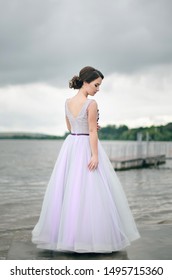 Beautiful Girl In Graduate Dress Near Water. Young Girl At The Prom With Gorgeous Hair In Purple Dress. Professional Make-up And Hairstyle, Stylish. Alone Girl Stands On A Pier Near Lake. Back View