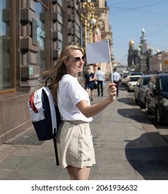A beautiful girl, a guide, blonde, in light clothes, shorts, with a white flag in her hand, in the daytime, invites tourists on a city tour. - Shutterstock ID 2061936632