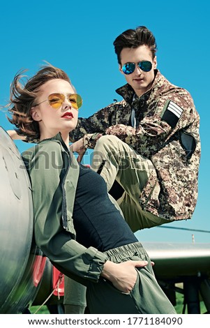 A beautiful girl and a handsome man pilots is posing next to a fighter jet at the airfield. Military aircraft. Military fashion.