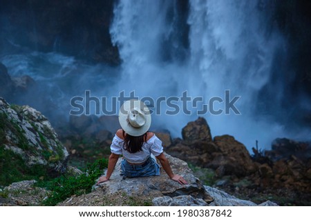 A beautiful girl in a hat sits on a stone and looks at the Kapusbashi waterfall in Turkey